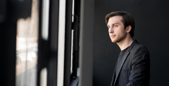 A young man with dark hair in a black shirt, looking out the window