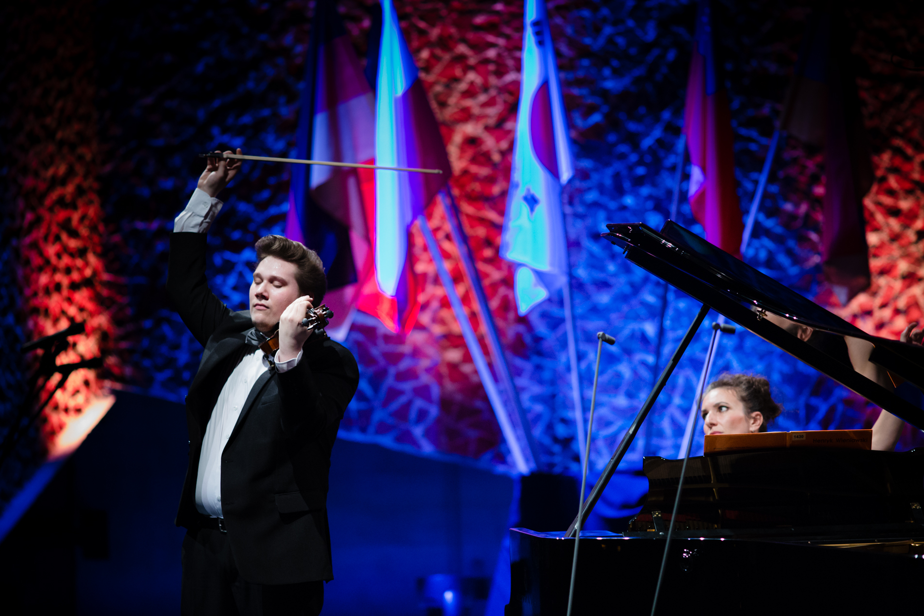 skrzypek, młody mężczyzna, uniesiona lewa ręka, trzymająca smyczek do góry.  w tle flagi różnych państw oraz pianista