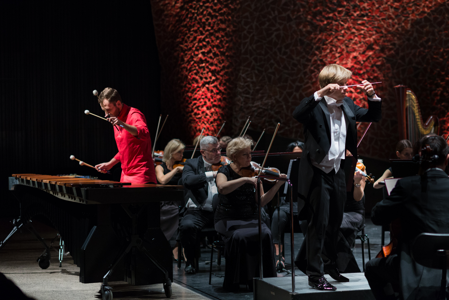 Orkiestra siedząca na scenie prowadzona przez blond dyrygenta, trzymającego batutę skierowaną do przodu. W roli solisty młody mężczyzna brunet w czerwonej koszuli grający na marimbie.