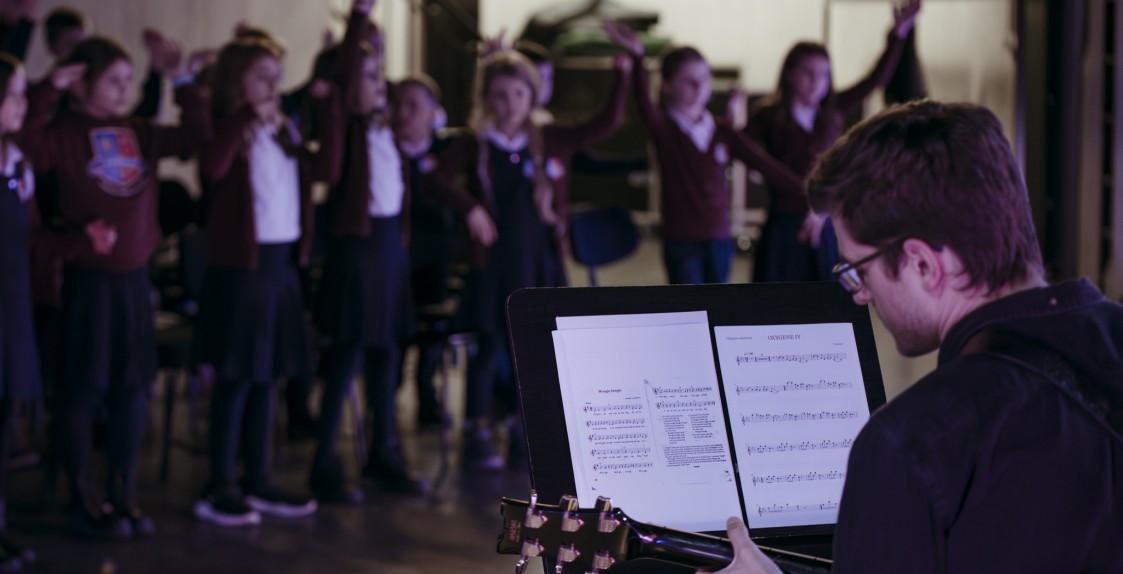 mężczyzna siedzący tyłem do fotografa grający na gitarze, przed nim pulpit z nutami a w tle skaczące dzieci