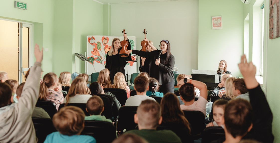 koncert w przedszkolu, dzieci zgłaszają się, prowadząca wskazuje na jedno z dzieci, dwie skrzypaczki pokazują skrzypce