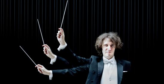 Mężczyzna trzymający w ręku batutę w czarnej marynarce i białej koszuli na czarnym tle