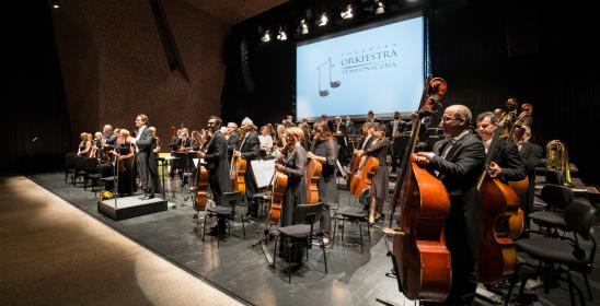 Orkiestra stoi po zakończeniu koncertu, w tle ekran z logotypem Toruńskiej Orkiestry Symfonicznej na białym tle