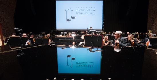 Orkiestra siedzi na scenie w tle widać ekran, na którym wyświetla się czarny logotyp na białej planszy. Dodatkowo ekran odbija się na klapie fortepianu.