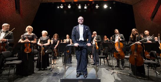 Zdjęcie orkiestry i dyrygenta stojących na scenie z instrumentami, witających Melomanów przed koncertem