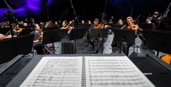 Orkiestra siedzi na scenie, na pierwszym planie otwarte materiały nutowe, z tyłu przyciemnione światło z fioletową poświatą.
