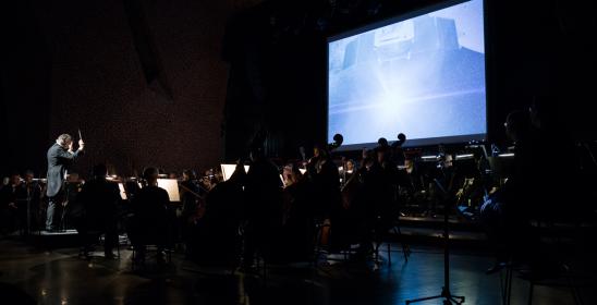 Orkiestra siedzi na scenie, w tle widać ekran, na którym wyświetlane są planety. Jest ciemno.