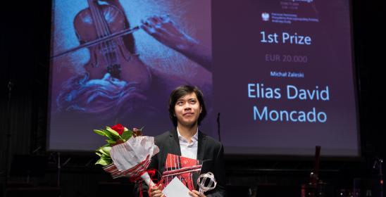 Młody chłopak z ciemnymi włosami trzyma bukiet kwiatów, z tyłu w tle wyświetla się prezentacja na ekranie z informacją, że zdobywcą pierwszej nagrody jest Elias David Moncado