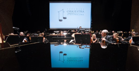 logotyp Toruńskiej Orkiestry Symfonicznej odbijający się na klapie fortepianu, w tle siedzący muzycy