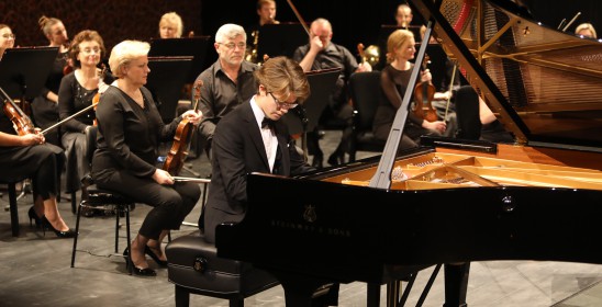 młody mężczyzna o blondowych włosach grający na fortepianie, za nim siedzą kobiety i mężczyzna ze skrzypcami w rękach
