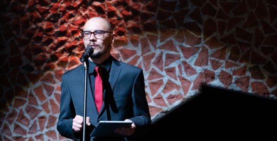 mężczyzna w ciemnym garniturze z czerwonym krawatem przemawia do mikrofonu