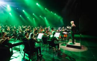 na scenie sali koncertowej w zielonym oświetleniu gra orkiestra symfoniczna na podeście stoi mężczyzna - dyrygent 