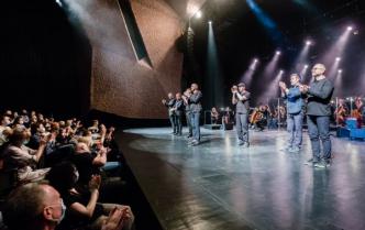 z lewej strony publiczność oklaskuje wykonawców koncertu stojących sześciu mężczyzn z zespołu Raz Dwa Trzy, w dali po prawej orkiestra symfoniczna