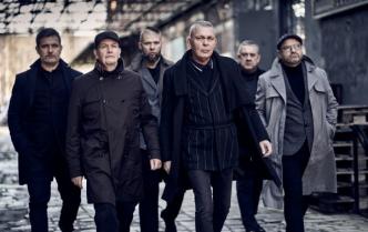 sześciu mężczyzn (zespół Raz, Dwa, Trzy) w płaszczach idą po bruku