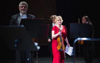 na scenie w śród pulpitów stoi kobieta z blond włosami w czerwonej sukni w prawej dłoni trzyma skrzypce w lewej kartki