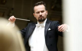 Uwertury i makarony mężczyzna Adam Banaszak -dyrygent w czrnym fraku z lekko uniesionymi rękami w prawej dłoni trzyma batutę drygencką