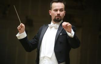 Uwertury i makarony mężczyzna Adam Banaszak -dyrygent w czrnym fraku z lekko uniesionymi rękami w prawej dłoni trzyma batutę drygencką