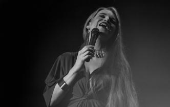 kobieta z długimi włosami śpiewa do mikrofonu, który trzyma w prawej dłoni, głowę ma lekko odchylona do tyłu. Zdjęcie zrobione w szarości