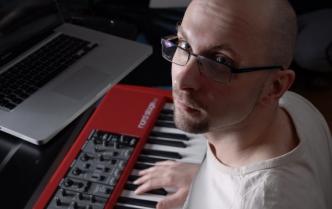 mężczyzna w okularach w białej koszulce patrzy w obiektyw dłoń trzyma na będącym przed nim keyboardzie