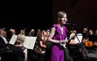 przy mikrofonie w fioletowym stroju stoi kobieta trzymając w dłoniach teczkę za nią siedzą muzycy 