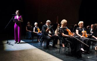 z prawej strony siedzą muzycy trzymając skrzypce z lewej przy mikrofonie stoi kobieta w fioletowym stroju