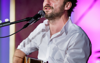 podczas koncertu mężczyzna w białej koszuli śpiewa i gra na gitarze, na jego twarzy widać zaangażowanie