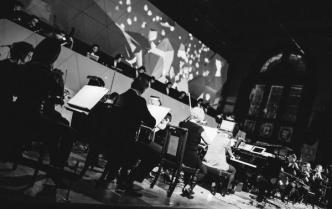 czarno-biała fotografia orkiestry, z przodu solista przy fortepianie, za orkiestrą DJe na podeście