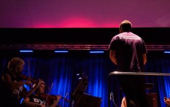 koncertmistrzynie obok dyrygent, wszyscy w fioletowych koszulkach, tlo podświetlone na różowo i niebiesko
