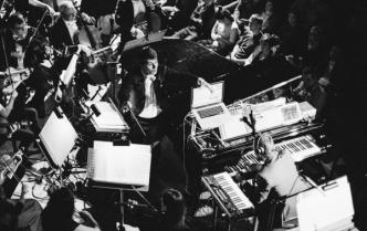 czarno- biała fotografia robiona z góry, na niej orkiestra wraz z dyrygentem i solistą siedzącym przy fortepianie z słuchawkami na uszach