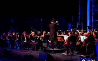 orkiestra siedzi na scenie, przed nimi dyrygent z uniesionymi rękoma do góry