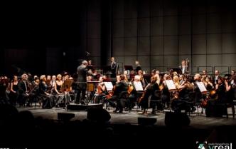 orkiestra gra na scenie z przodu dyrygent, w tle ściana z muszli koncertowej