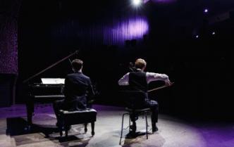 Aleksander Dębicz siedzi przy fortepianie  obok siedzi Marcin Zdunik  grając na wiolonczeli, zdjęcie zrobione z tyłu sceny dlatego widać plecy i tył głowy mężczyzn oraz klawiaturę fortepianu