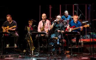 sześciu mężczyzn gra i śpiewa siedząc na scenie