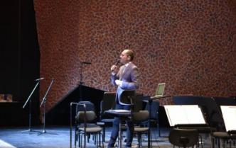 na scenie stoją ustawione krzesła i pulpity do nut przed nimi stoi trzymając w ręce mikrofon prowadzący koncert Adam Suprynowicz