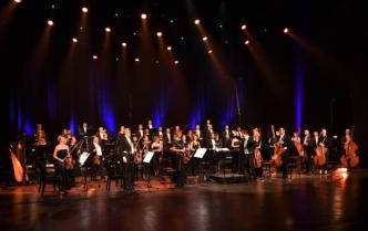 na scenie stoi orkiestra -kobiety i mężczyźni trzymający instrumenty przed nimi stojący bokiem  dyrygent - Adam Banaszak