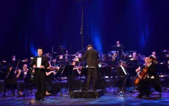 na scenie śpiewa Łukasz Gaj - tenor za nim tyłem stoi dyrygent dyrygując orkiestrą grającą na instrumentach