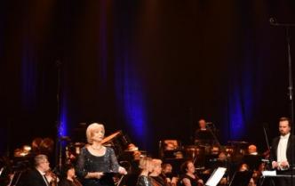 na scenie prezenterka Urszula Guźlecka za nią siedząca na krzesłach orkiestra i stojący przodem dyrygent