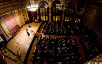para skrzypków na oświetlonej scenie, przed sceną publicznośc w fotelach, zdjęcie z góry
