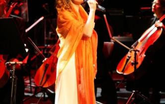 kobieta z długimi włosami w kremowej sukni i z pomarańczowym szalem na ramionach stoi na scenie a mikrofonem w dłoni