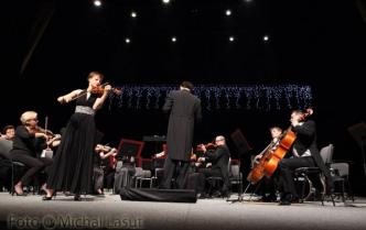 orkiestra siedzi na scenie na czarnym tle w nastrojowym świetle, z przodu solistka gra na skrzypcach  w czarnej sukni