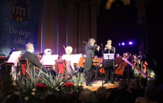 orkiestra siedzi na scenie w nastrojowym fioletowym świetle, z przodu soliści, skrzypek i trębacz