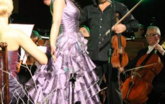 śpiewająca kobieta w fioletowej sukni, za nią przyglądający się skrzypek, wokół grający muzycy orkiestrowi