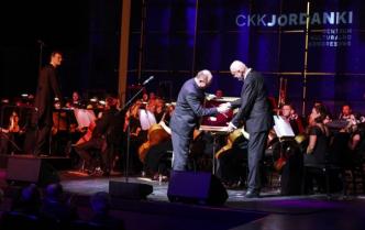 marszałek i prezydent Torunia ściskają swe dłonie na scenie, za nimi orkiestra