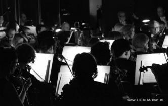 czarno-biała fotografia pokazująca kontury muzyków z instrumentami na tle ich podświetlonych nut