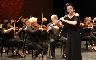 kobieta stojąc gra na flecie poprzecznym za nią siedzący muzycy kobiety i mężczyźni grają na skrzypcach