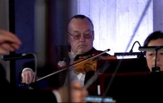 mężczyzna siedząc gra na skrzypcach przed nim z lewej ręce z pałeczką dyrygencką, po prawej przed pulpitem twarz kobiety