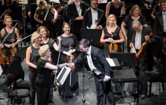 na tle stojących uśmiechniętych muzyków Toruńskiej orkiestry symfonicznej na scenie dyrygent przybija żółwika z Panią koncertmistrz