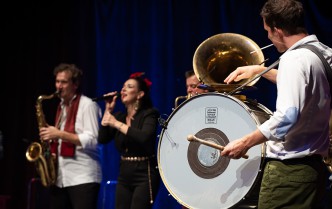 a man playing the drum, in the background a woman singing into the microphone and a man playing the saxophone