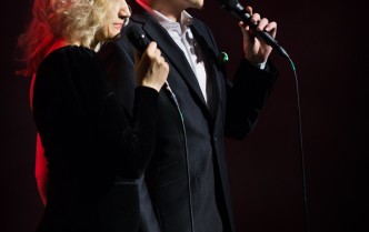 obok siebie stoją bląd włosa kobieta i mężczyzna z ciemnymi włosami, ubrani w ciemne stroje w dłoniach trzymają mikrofony
