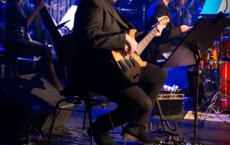 mężczyzna siedząc gra na gitarze basowej w tle muzycy orkiestry symfonicznej